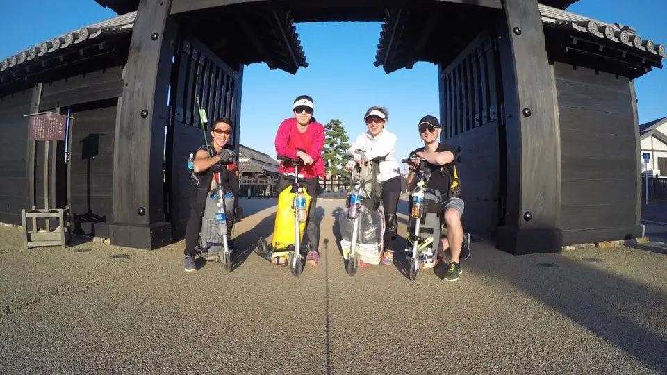 Team of four kick scooter adventurers posing with their loaded kick scooters at a traditional Japanese gate structure, ready for their cross-country journey