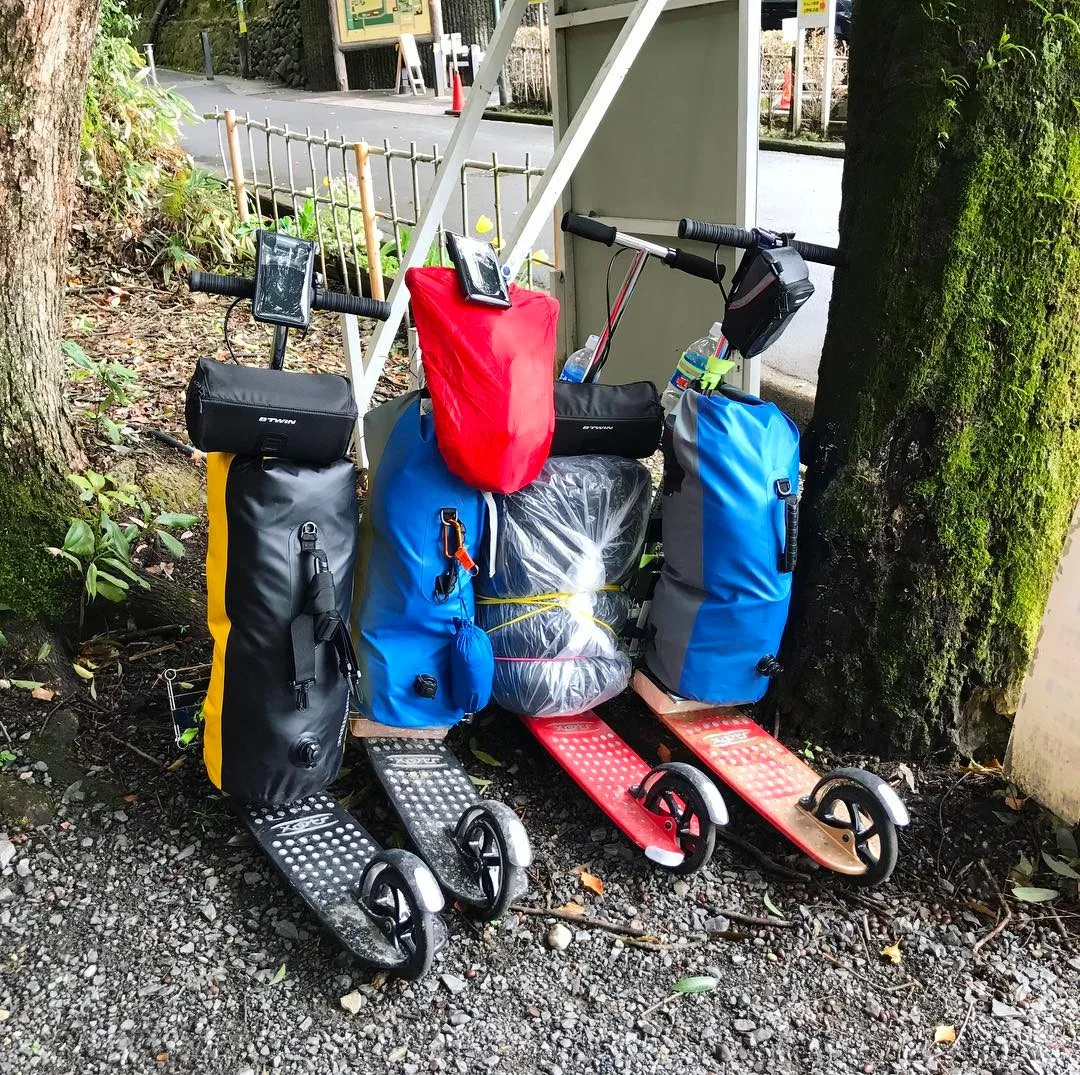 Four kick scooters with travel gear including panniers, dry bags, and camping equipment, demonstrating how kick scooters are used for multi-day travel adventures